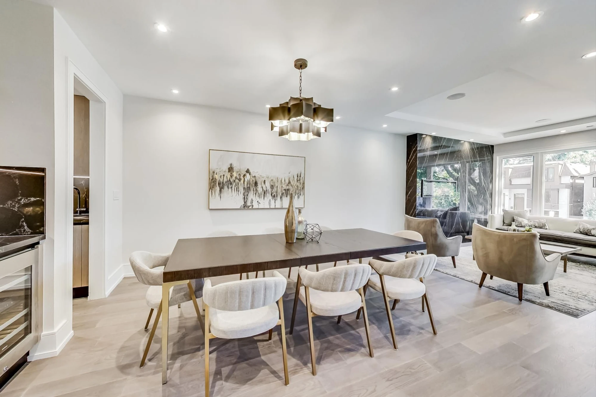 A bright dining room with a dark wood table, cream chairs and a brass chandelier, opening to a living room with a marble fireplace wall