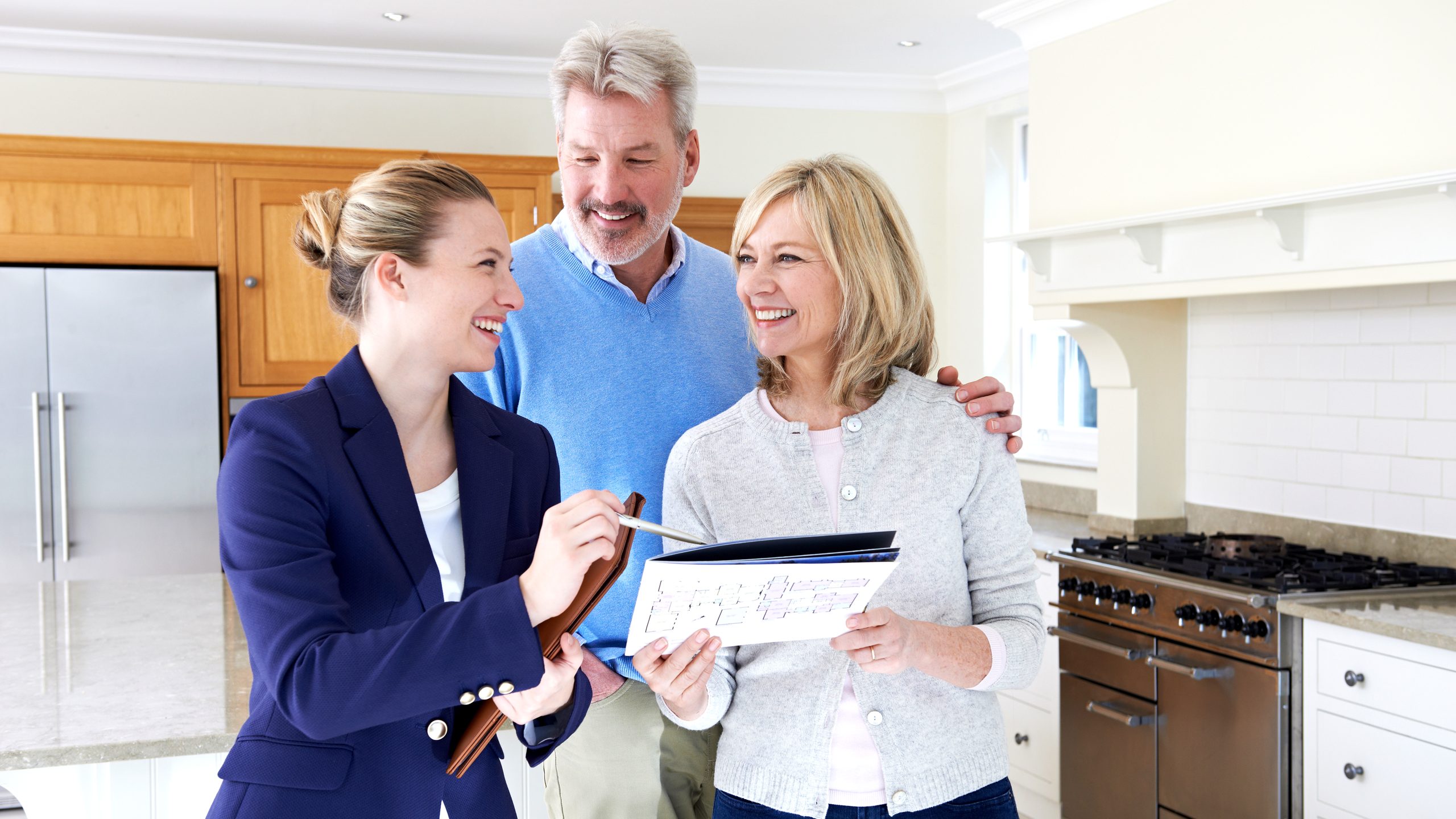 A real estate adviser goes over a floor plan with an older couple in a bright traditional kitchen