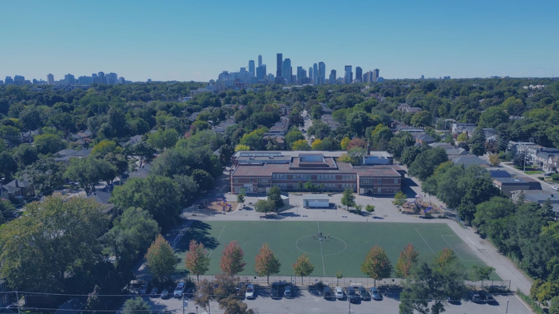 Aerial view of a school and sports field among tree-lined streets, with towers in the distance