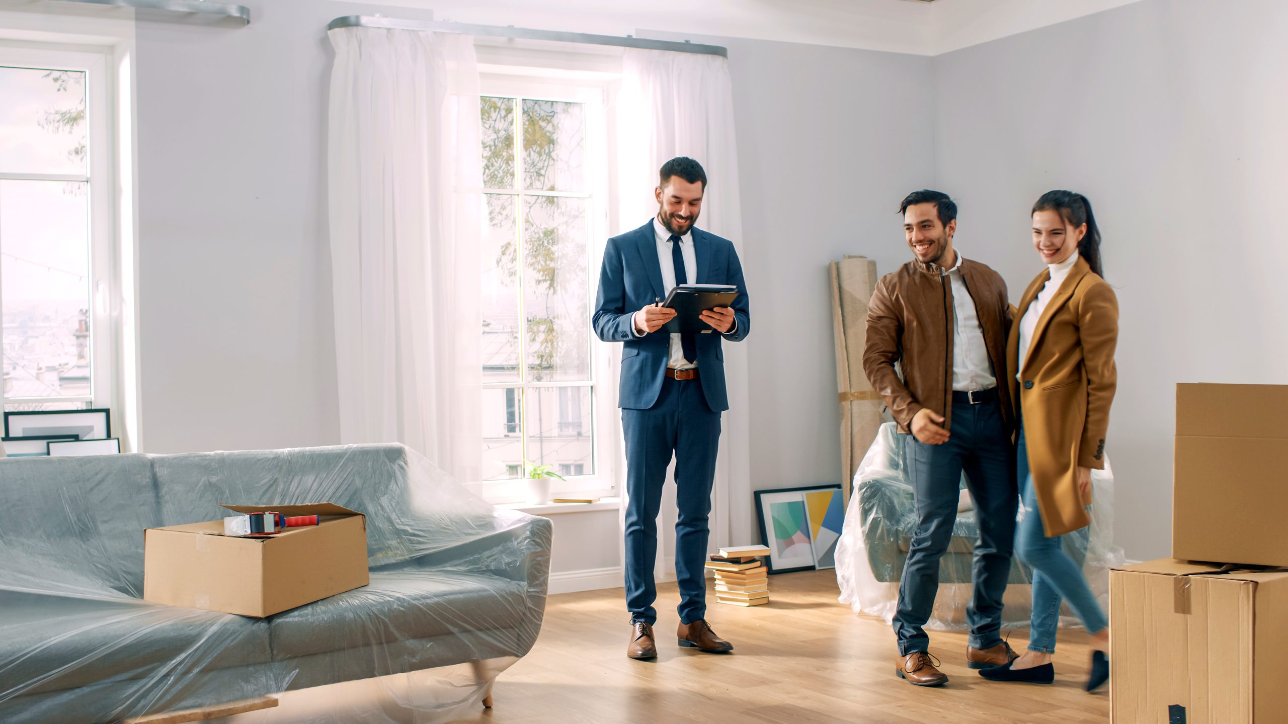 A young couple in winter coats looks around their new home with a real estate agent, moving boxes beside them