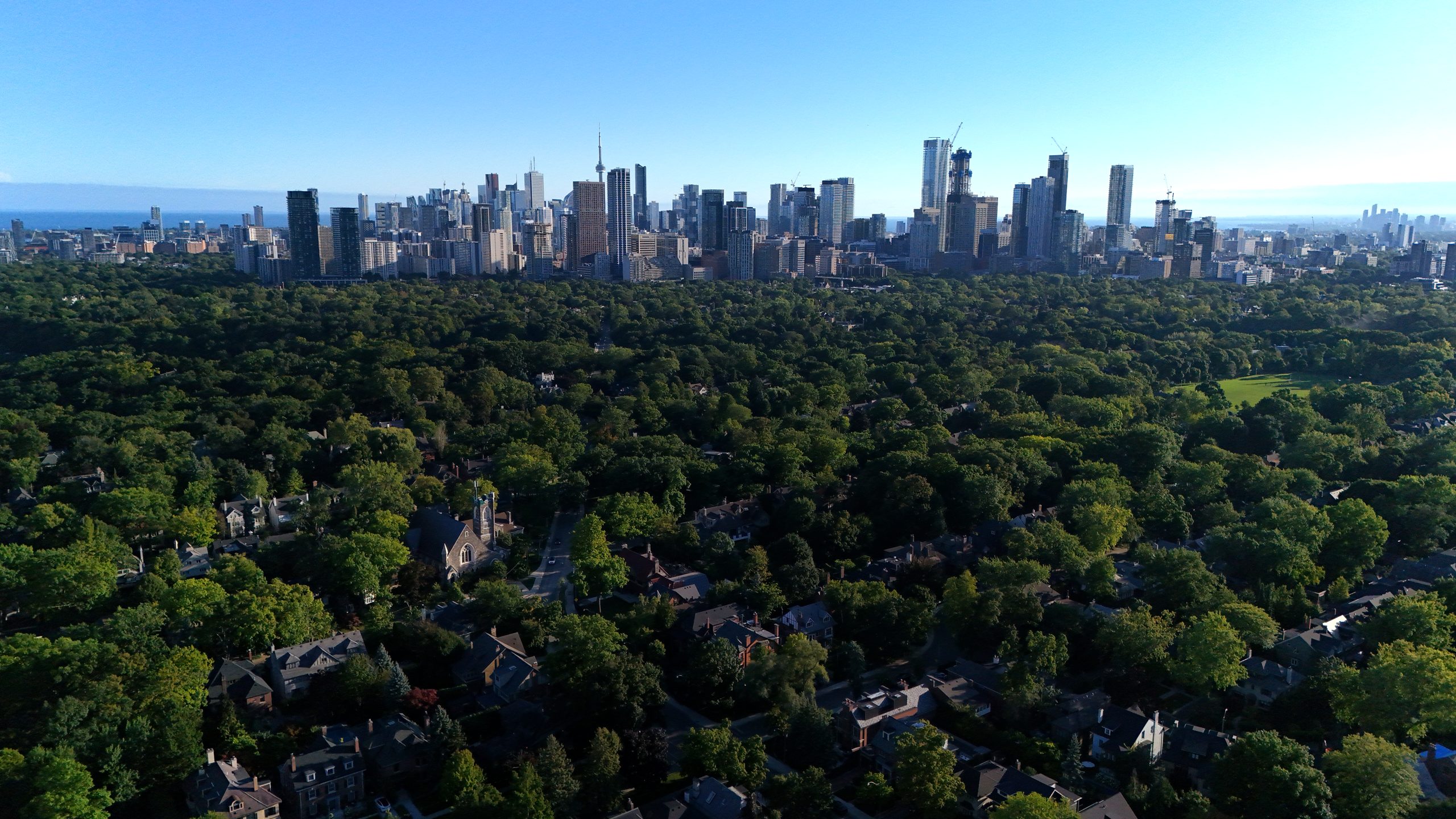 Drone view south over the Rosedale tree canopy to the downtown Toronto skyline and CN Tower at golden hour.